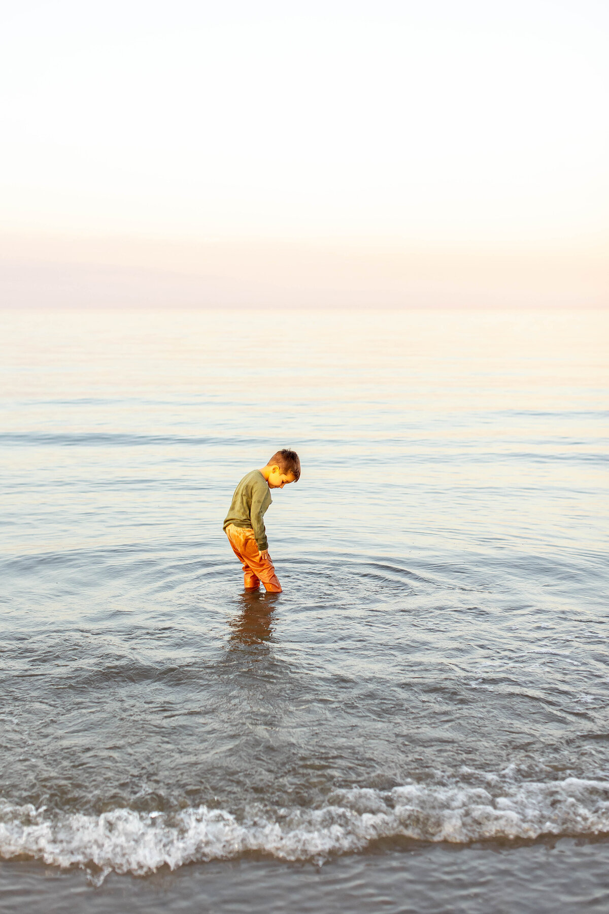 A young boy explores the quiet ocean shallows at sunset, captivated by the golden ripples dancing around in Virginia Beach