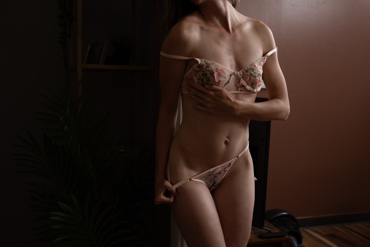 A woman in pink lace underwear pulls down her bottoms while standing in a New England Boudoir studio