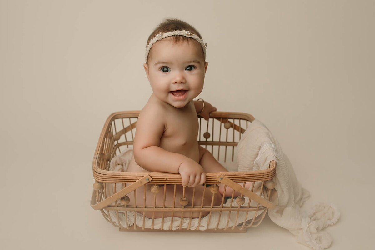 a six month old girl sitting sideways in a lovely basket with a white fabric draped over the sides