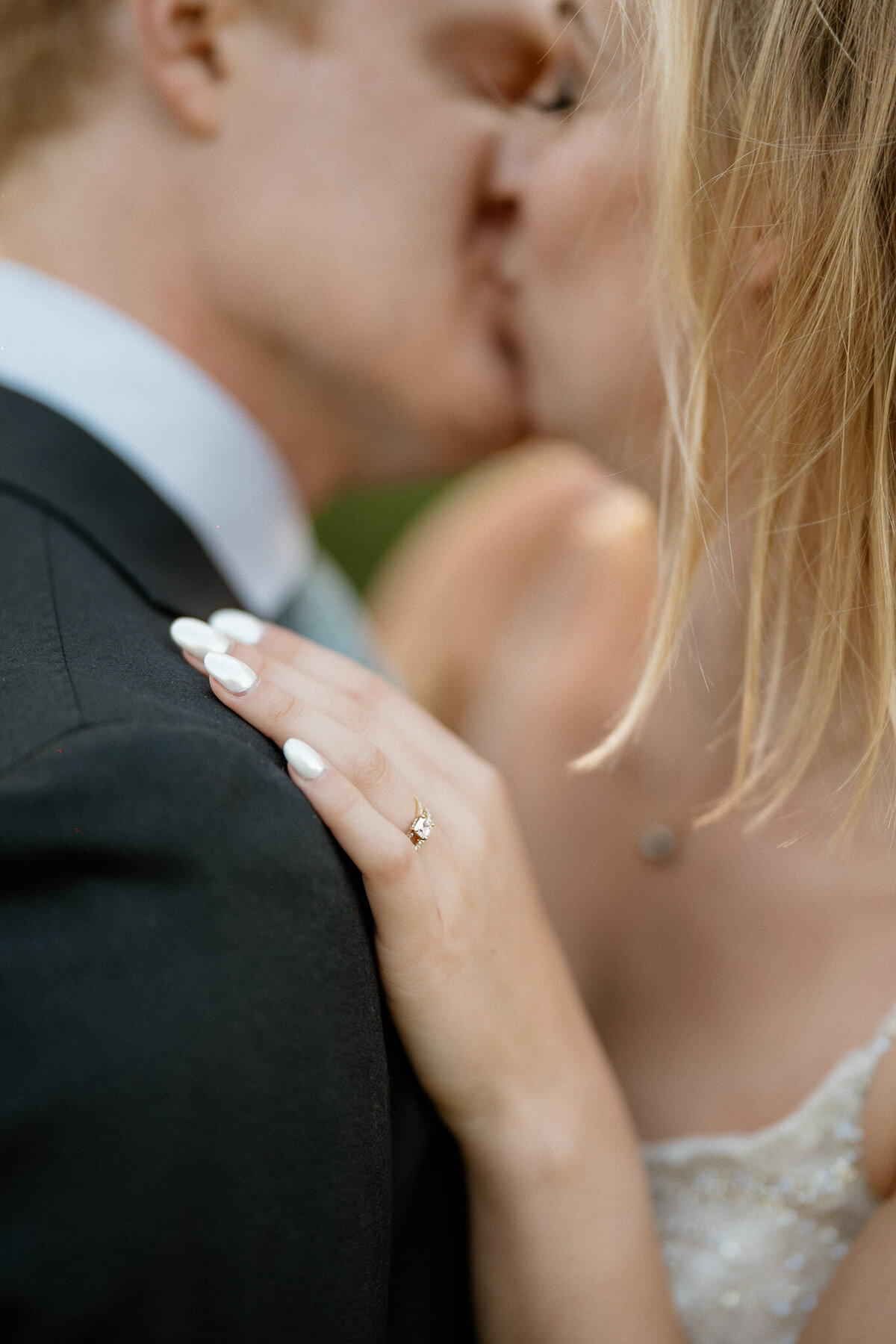 A bride and groom kiss while the bride rests her hand with diamond ring on the shoulder of the room