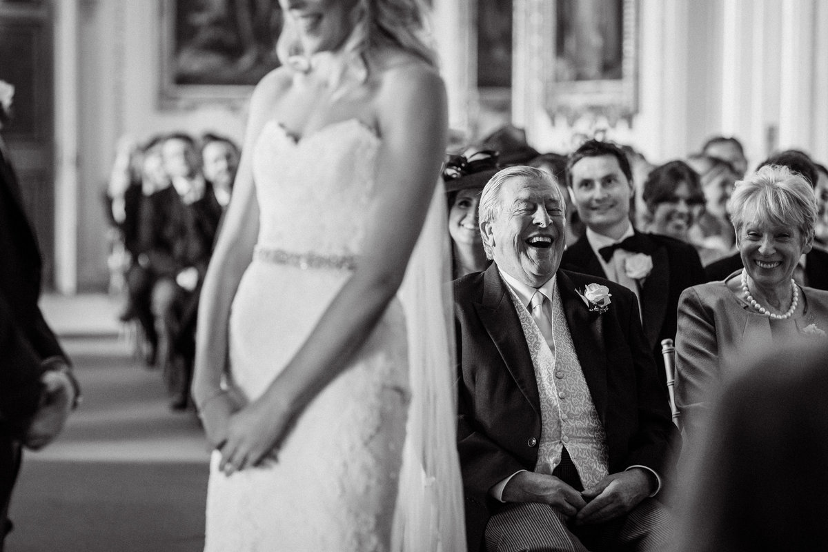 Black and white wedding photography at Goodwood House of the bride blurred in the foreground and the father of the bride behind laughing with emotion during the ceremony