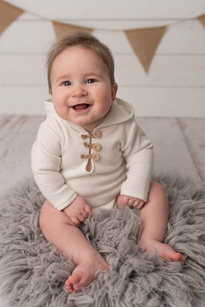 Six month old boy sitting on rug, smiling at the camera in white romper