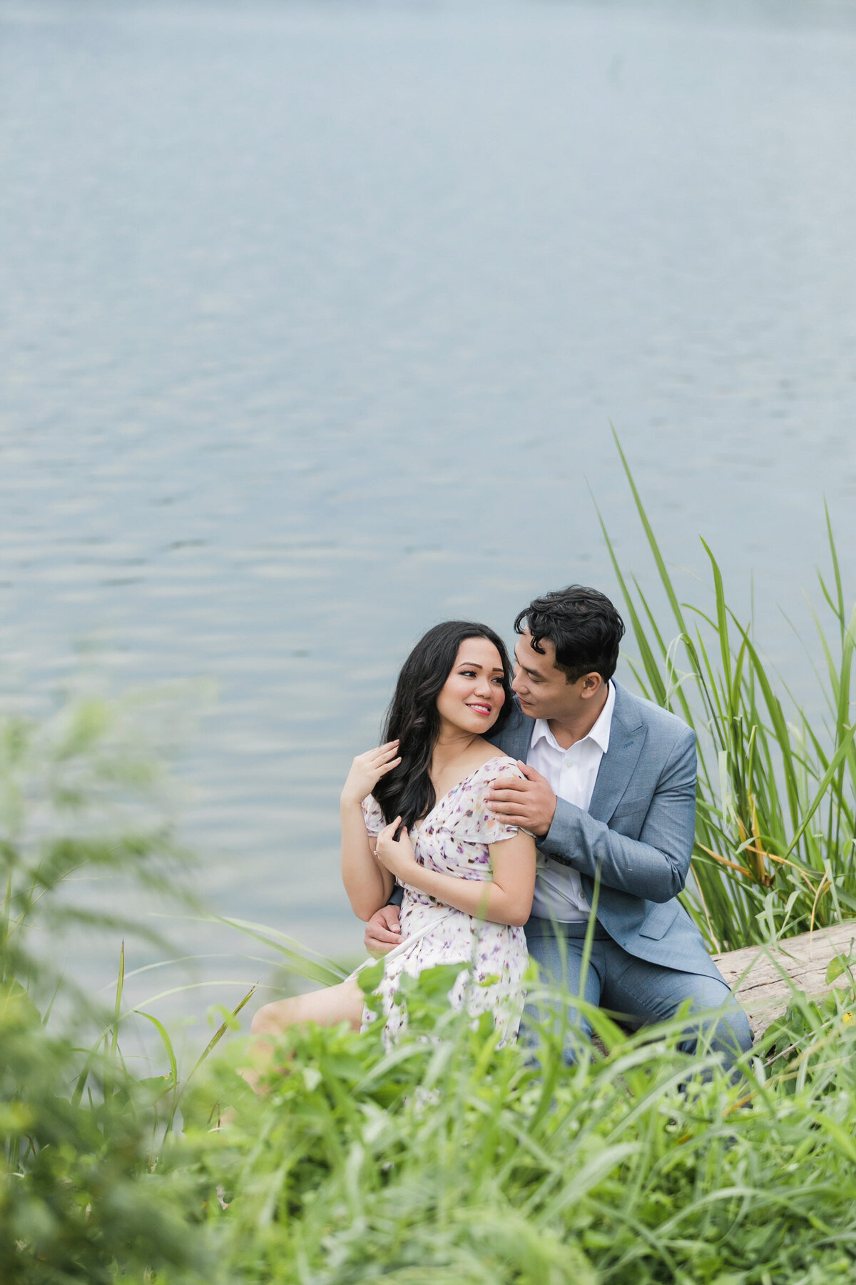 Couple seated at White Rock Lake during their engagement session, with the water behind them, looking at each other in a romantic moment.