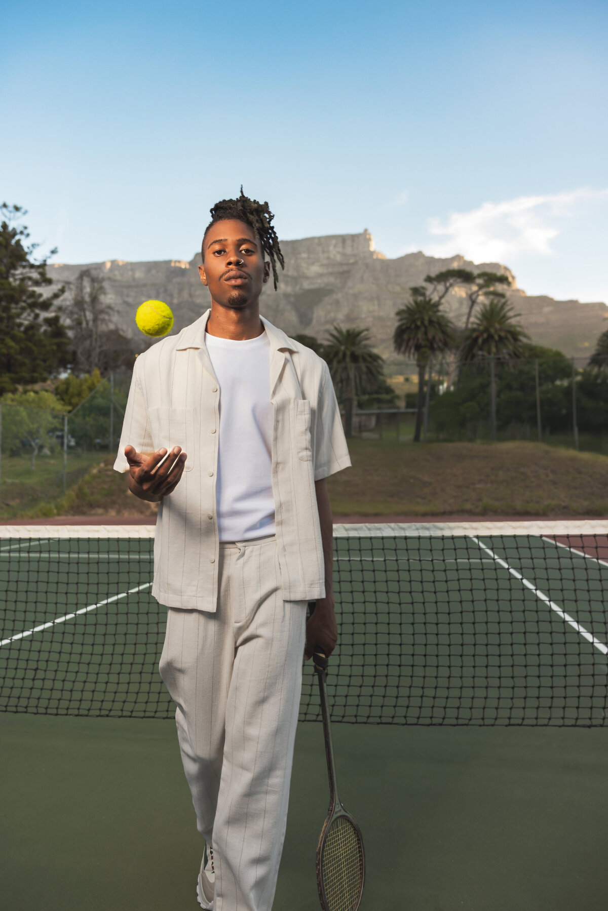 Stylish man in a cream-toned outfit tossing a tennis ball on an outdoor court, with Table Mountain and palm trees in the background.