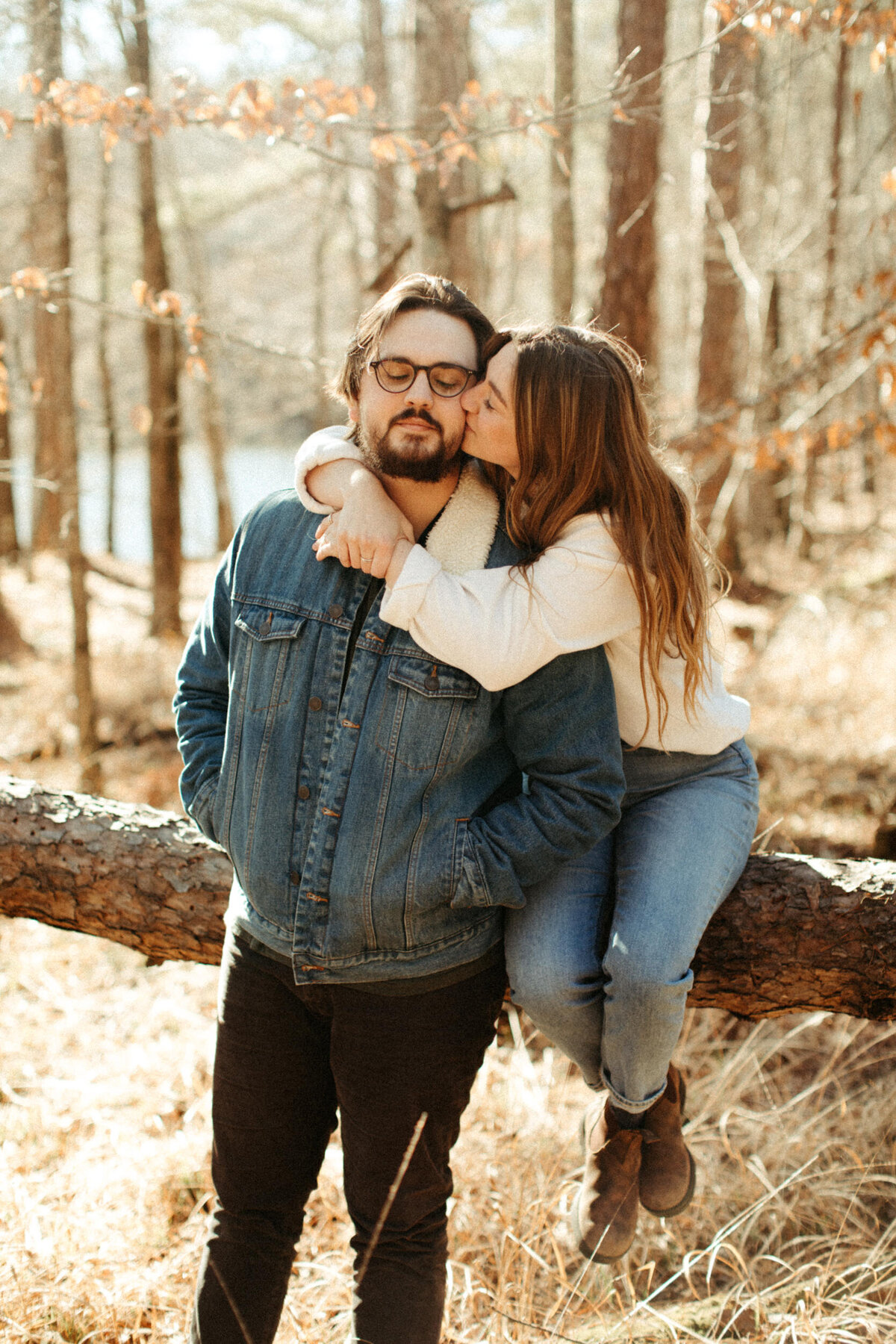 A girl is sitting on a fallen tree log in the woods while she hugs and kisses her fiancé who is standing next to her and wearing a denim jacket.