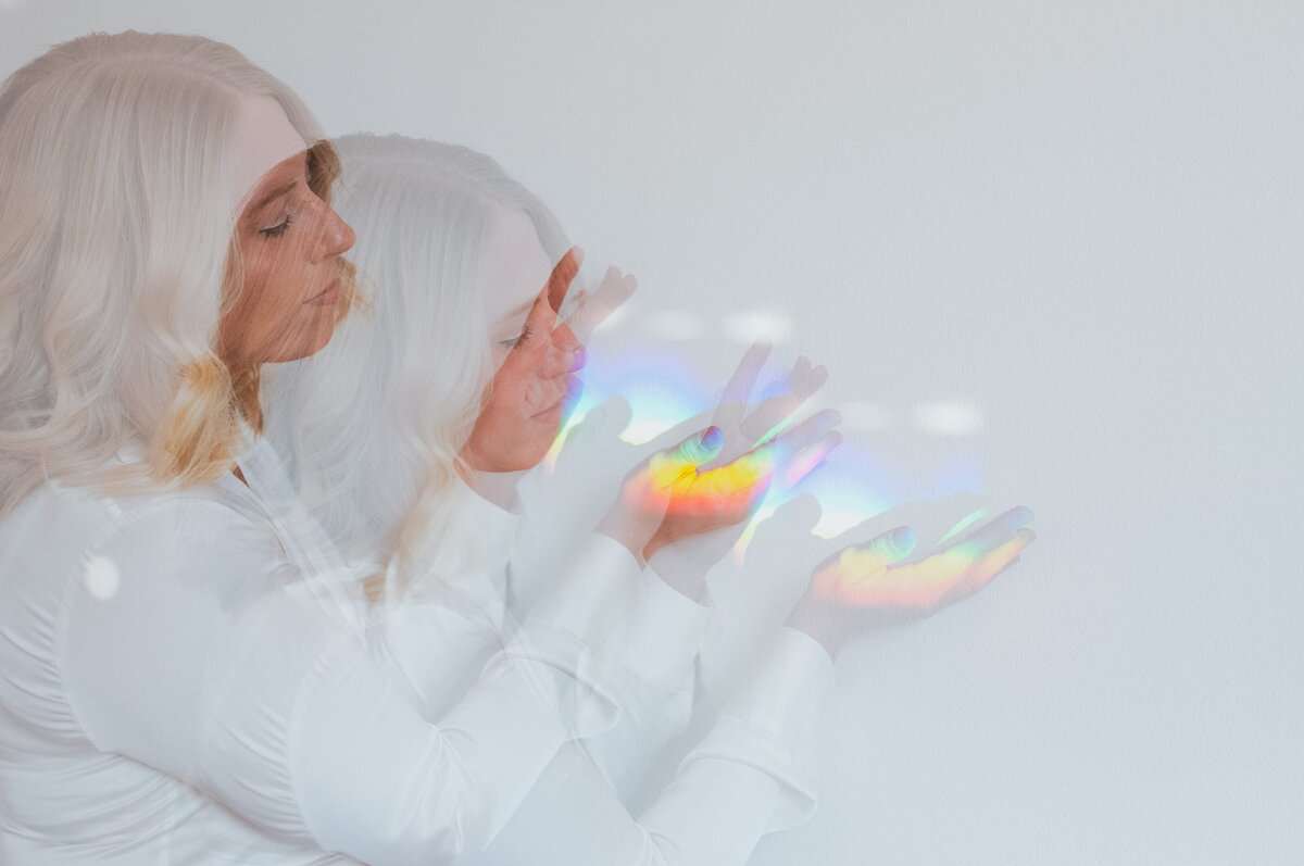 Double exposure of woman with blonde, wavy hair and silk, white top with rainbow and disco ball reflections on her as she reaches both hands out to play with reflections.