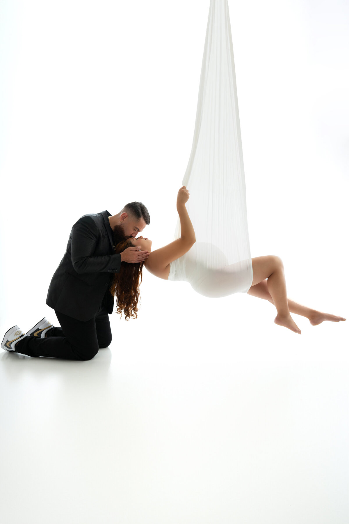 Partner gently touches mom head as she is suspended in the air by a white flowy fabric. 