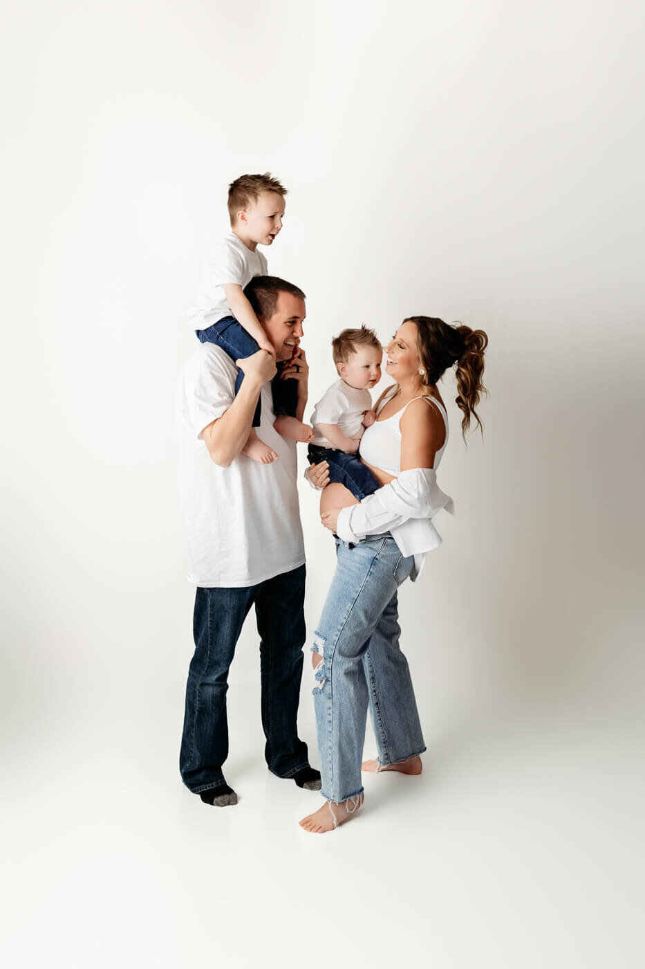 A family of four smiles together in a studio, perfect for maternity portraits. The father holds a young boy on his shoulders, while the mother—captured by a Bloomington maternity photographer—holds another young boy; all wear white tops and jeans.