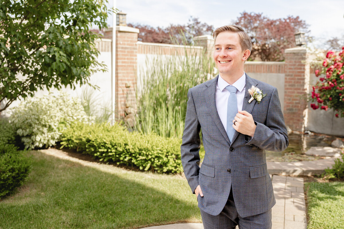 A groom in a gray suit, boutonnière pinned, smiles confidently in a beautifully landscaped garden.