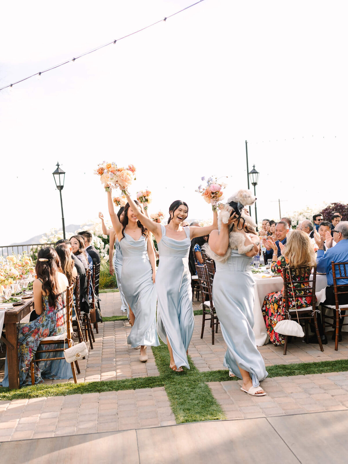 Bridesmaids in flowing blue dresses joyfully walk down an outdoor aisle, holding floral bouquets, as guests celebrate at elegant tables.