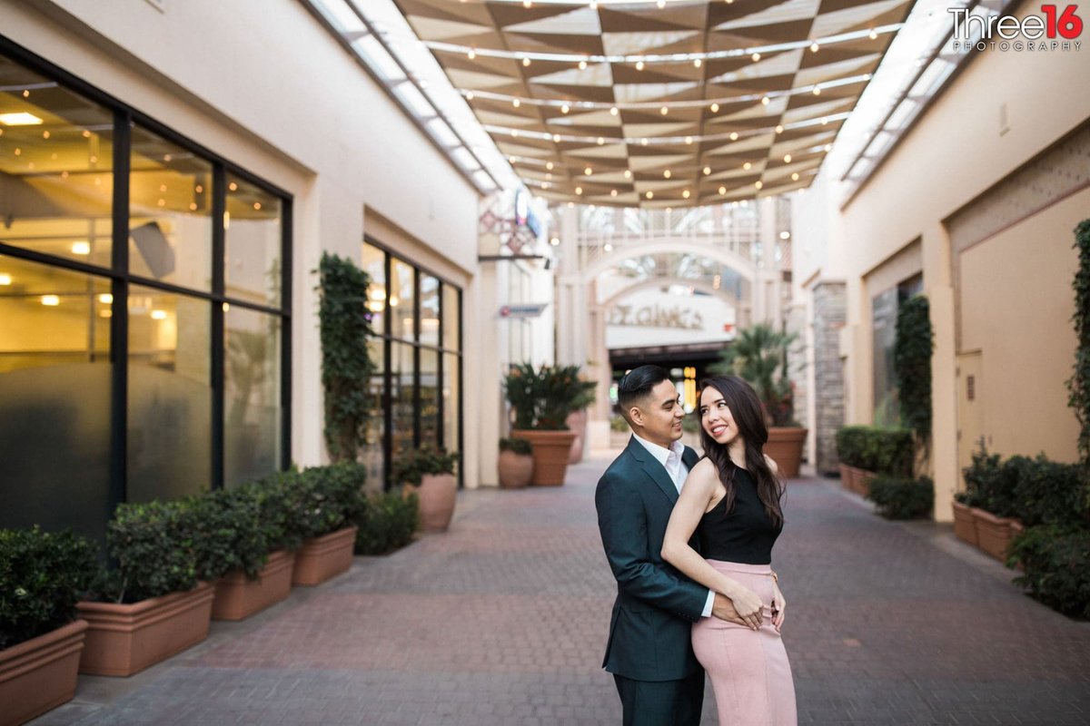 Bride to be looks back at her Groom as he holds her in the walkways of the Irvine Spectrum