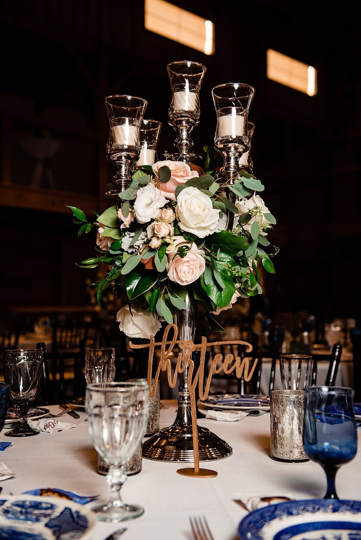 Tall silver candelabra centerpiece at Sycamore Farms wedding reception. Wedding reception table set with white floral centerpiece with silver candelabra, blue mismatched vintage china, blue and clear vintage glass water goblets and a white table cloth.