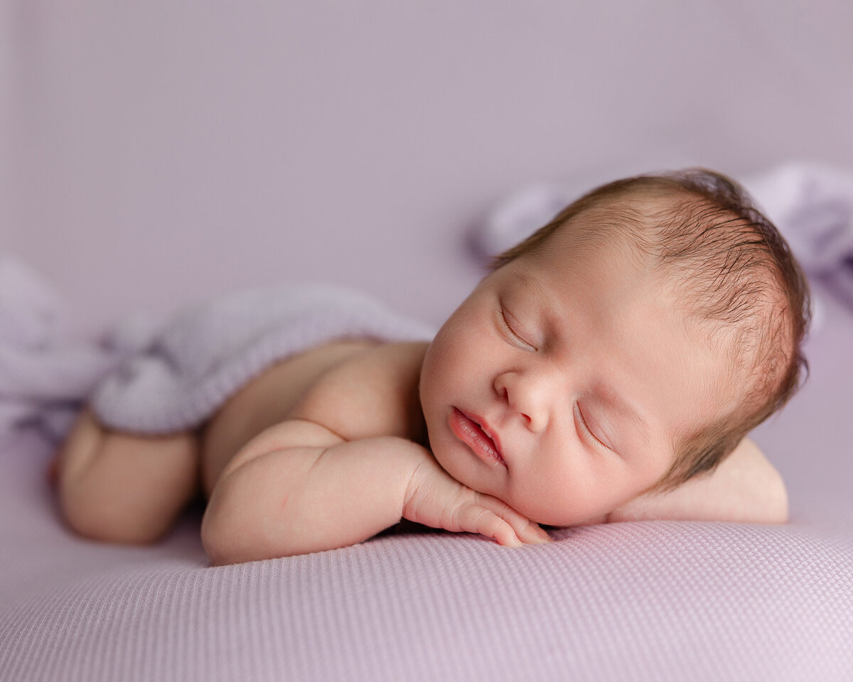 Newborn baby boy peacefully sleeping on his side, wrapped in soft lavender tones, with a relaxed pose and gentle expression. Captured during his Inland Empire newborn session by Kaitlyn Dawn Photography