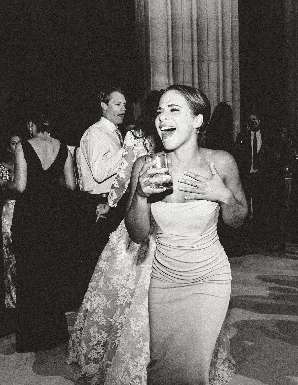 At Washington National Cathedral in Washington D.C., the photo was taken by photographer Twah Dougherty. It beautifully captures a woman in a strapless dress, holding a drink, laughing heartily at a formal event. The background features other attendees in elegant evening attire engaged in conversation and dancing, with tall columns enhancing the sophisticated setting of the scene. To further enhance the event, you can consider adding Current Films as videographers to your information for capturing the lively moments in motion. The black and white tones in the photo add a classic and timeless charm to the overall composition.