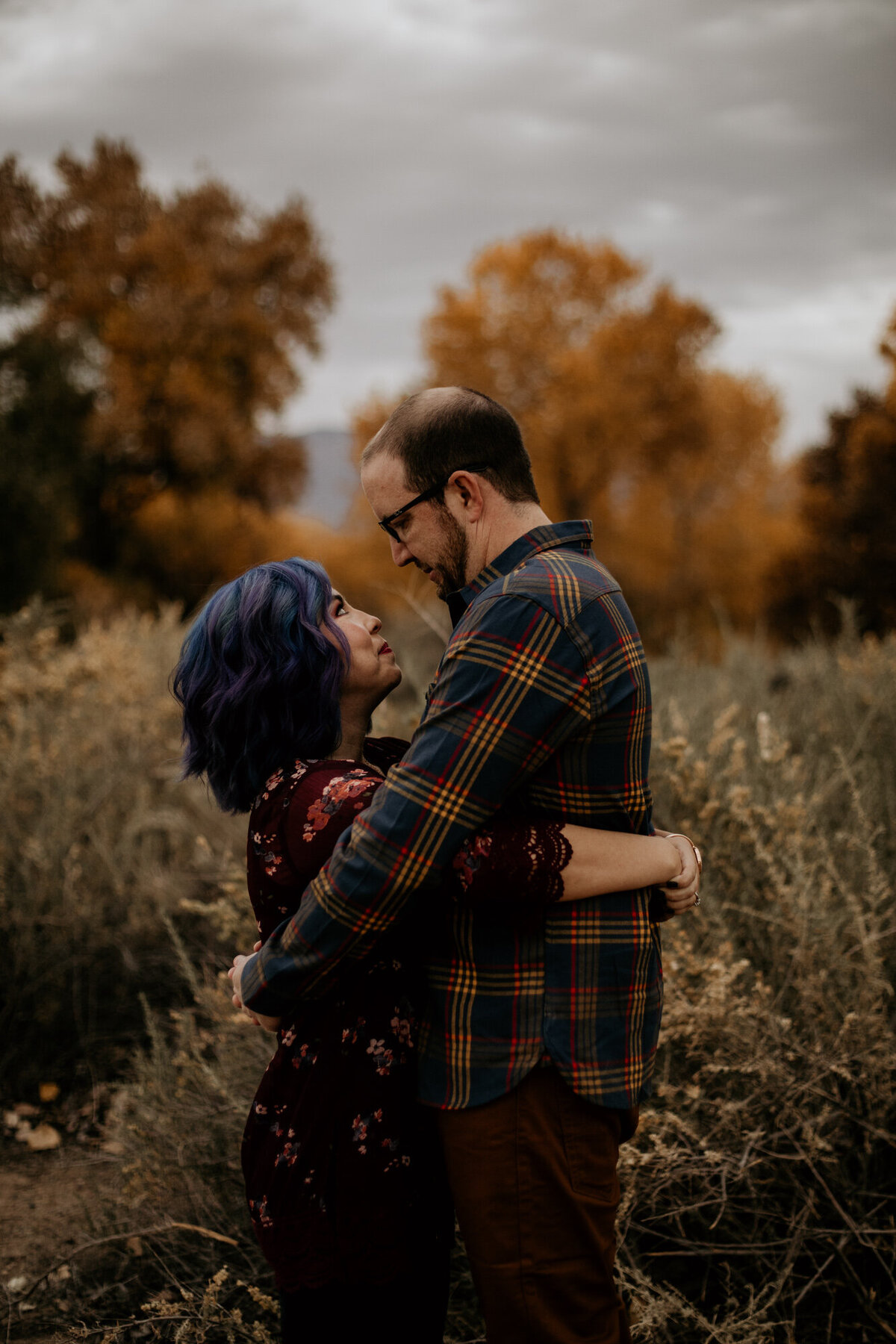 blue haired woman hugging man in front of fall trees