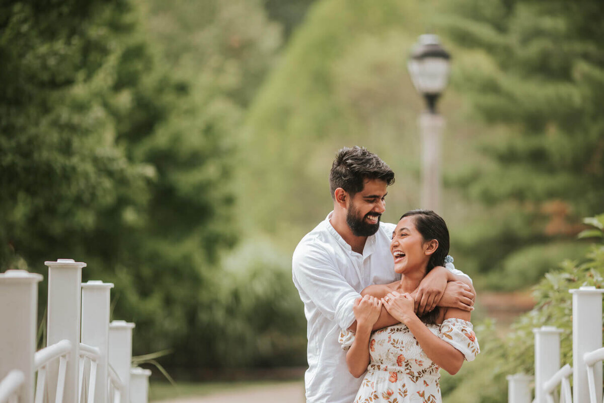 A joyful couple embraces on a charming bridge surrounded by lush greenery in Hamilton Square, NJ.