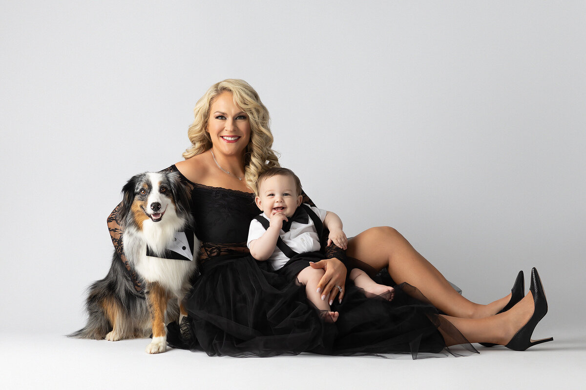 A stylish mom in a black lace gown poses with her baby boy in a tuxedo outfit and their well-groomed dog in a matching bow tie, captured by a Dallas milestone photographer.