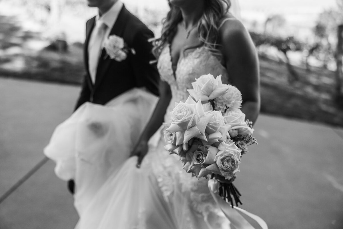 Black and white close up of bride walking with her bouquet. Her husband helps carry the train of her wedding dress
