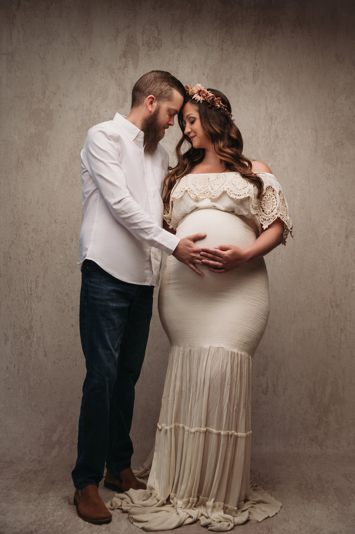 dad to be holding pregnant brunette woman's baby bump who is wearing a boho cream maternity gown and touching foreheads with their eyes closed