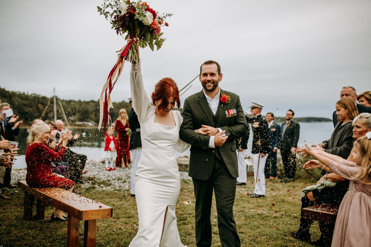 couple exits from their wedding ceremony off the coast of South Bristol Maine, flower petals are thrown by guests and bride waives hand in the air with bouquet to celebrate