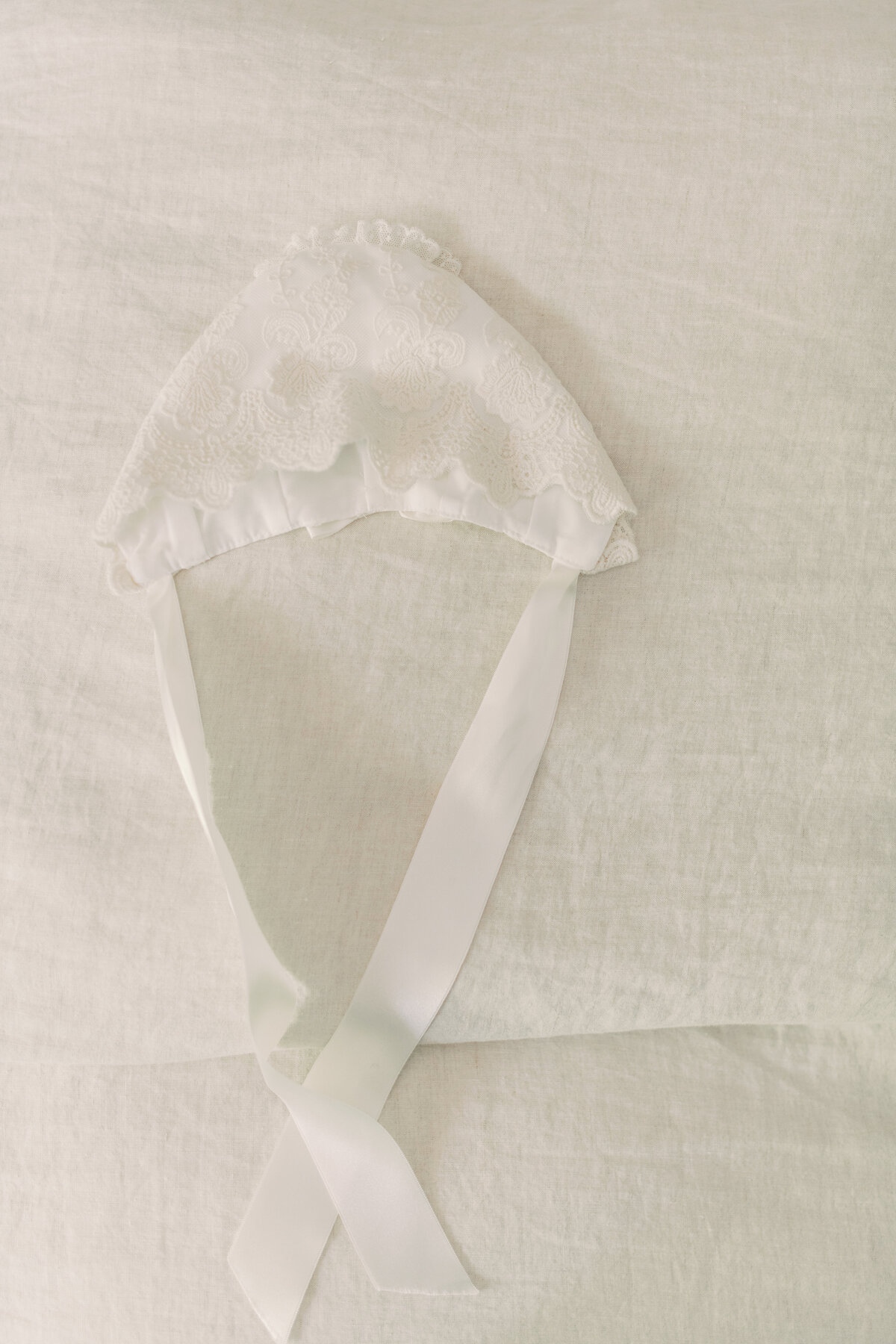 Elegant detail of baptism bonnet ribbon held by baby, surrounded by soft textures and ivory tones.
