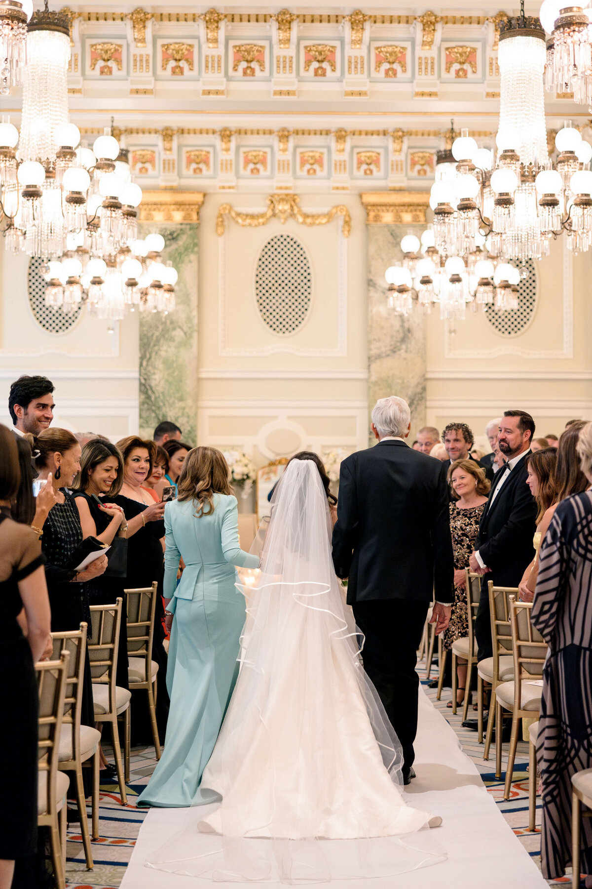A bride, wearing a white gown and veil, walks down the aisle with two people in formal attire. Guests sit and stand, watching in a grand, elegantly decorated hall with chandeliers and ornate walls.