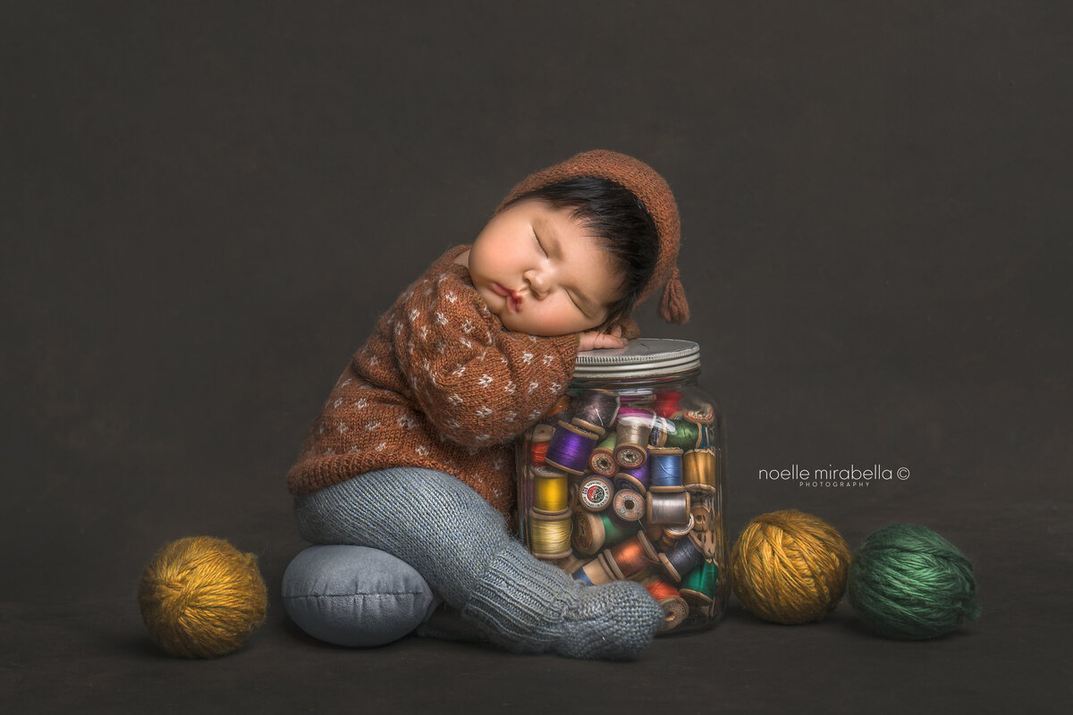 Newborn baby in handmade knits sleeping on glass jar of vintage thread spools.