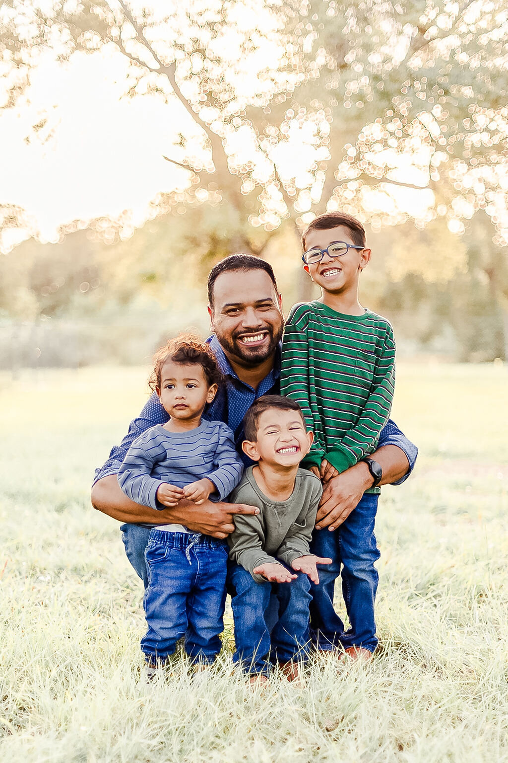 a joyful outdoor portrait of a father and his three young sons. The father is kneeling on the grass with a big smile, hugging his children closely. One child, wearing a green striped shirt and glasses, stands next to him, smiling brightly. Another child, in a green shirt, crouches down and grins widely with his eyes closed. The youngest child, dressed in a blue striped shirt, stands calmly with a more serious expression. The background is a sunny park with tall trees and a soft golden glow from the setting sun.