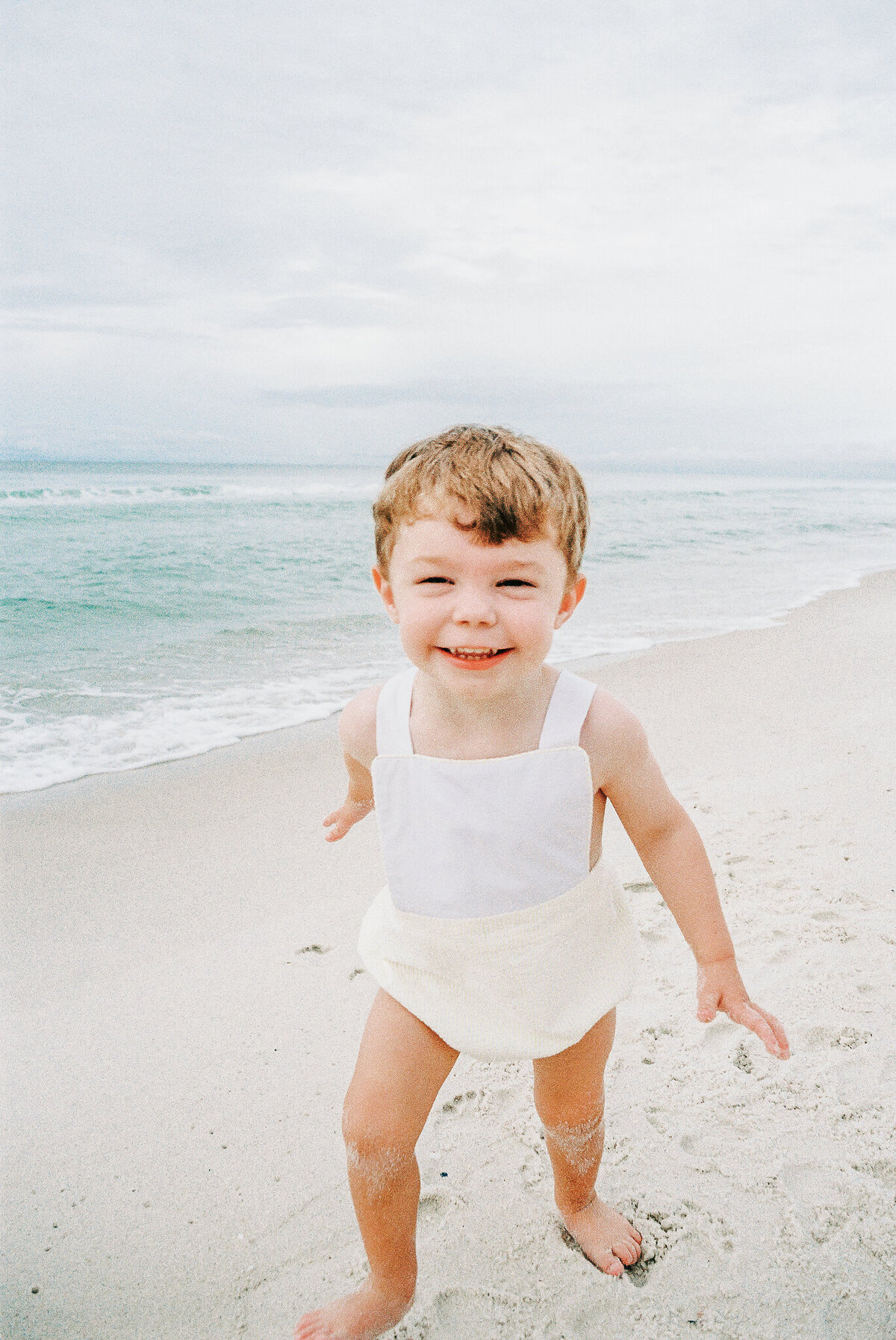 A joyful young boy runs barefoot along the sandy beach, his face beaming with happiness. Dressed in a classic white and yellow romper, he plays near the gentle ocean waves, capturing the carefree essence of childhood in a light and airy family portrait by a Birmingham family photographer.