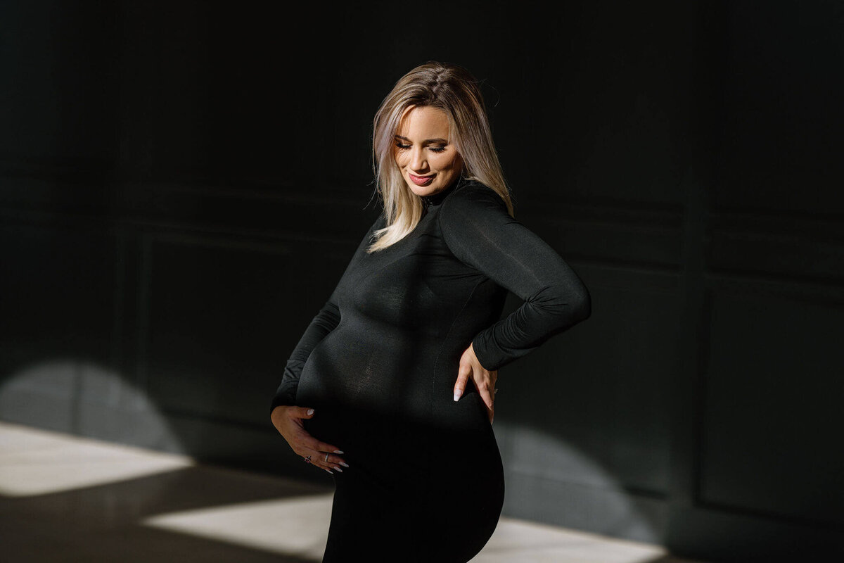 fine art maternity portrait of expecting mother in black dress standing in window light
