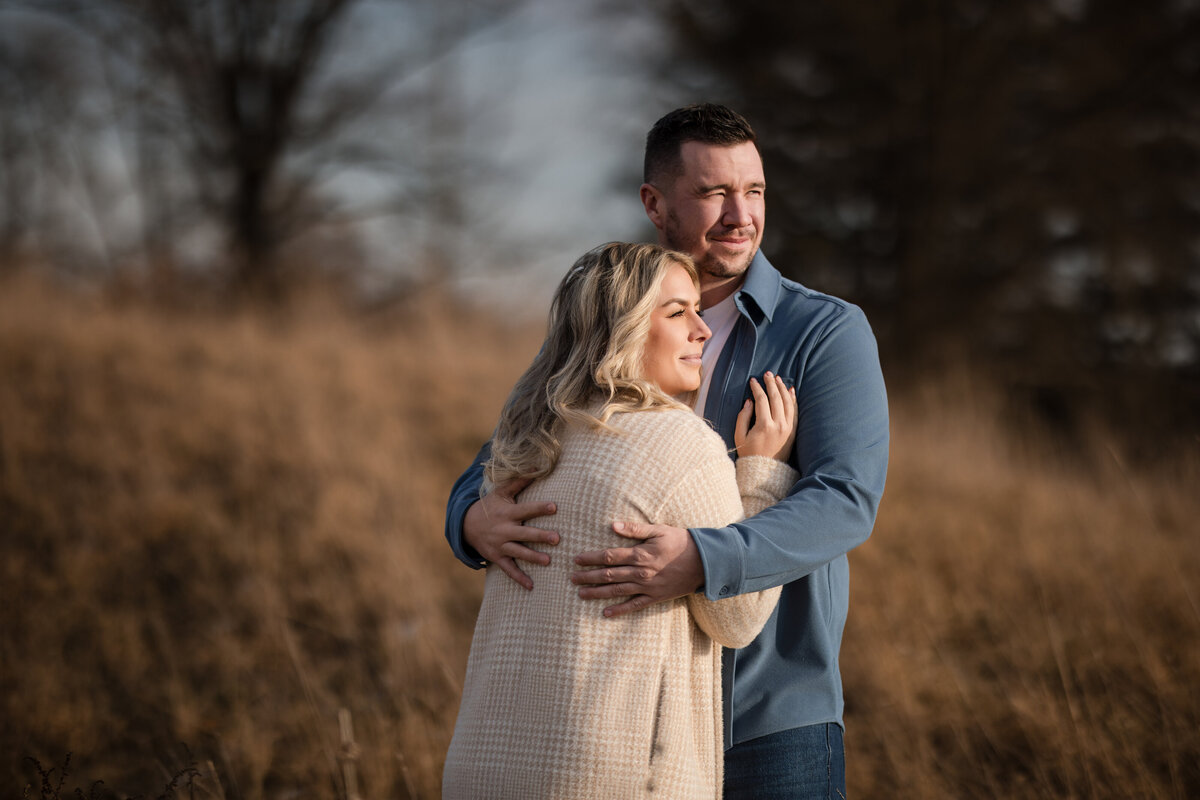 Couple embracing in a golden field of tall grass, with soft afternoon light casting a warm glow—this image is part of a series of Terrain engagement photos, capturing a quiet, natural moment that reflects the season and setting.
