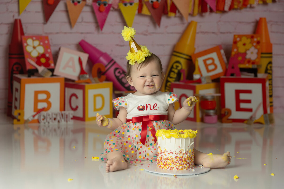 A toddler girl in a crayon themed studio sits on the floor smiling behind a small cake