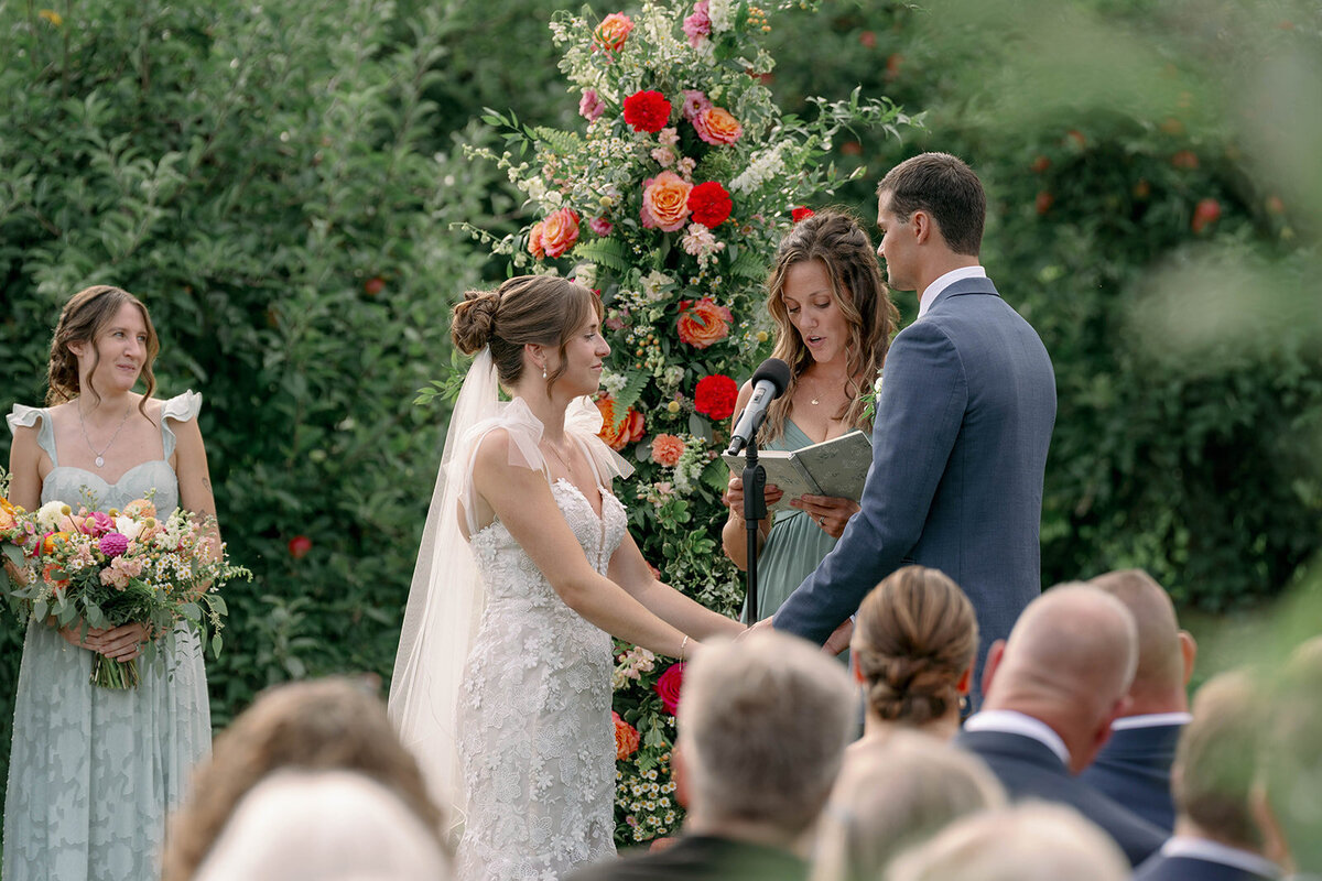 Bride and groom holding hands during their orchard ceremony at The Cherry Barn in Frankfort, Michigan, captured during their emotional vow exchange.