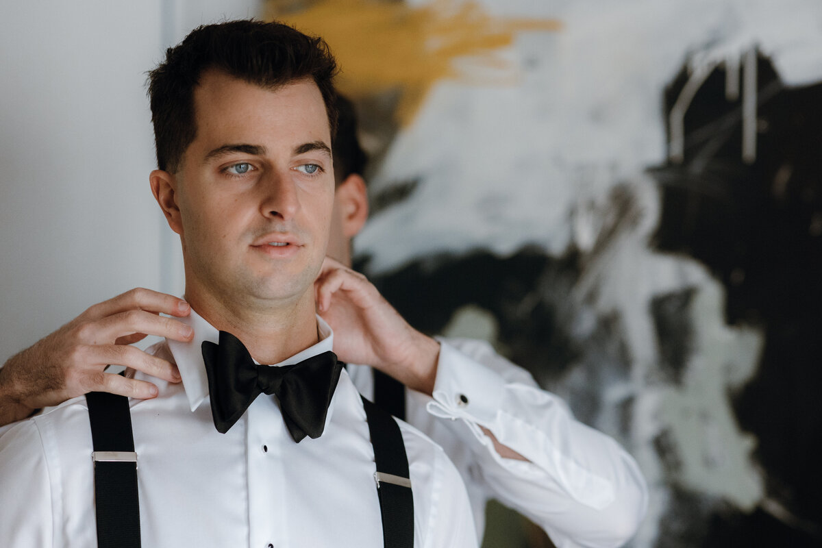 A groom having his collar and bowtie fixed by his partner behind him.