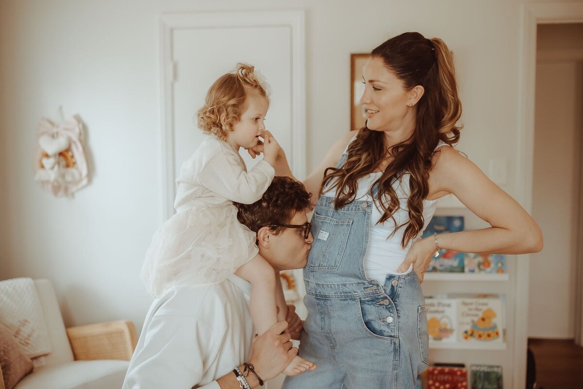A woman in overalls beams as a child perches on a man’s shoulders, with the man playfully kissing her belly. Books line the background, capturing a heartwarming moment perfect for a Macon maternity photographer.