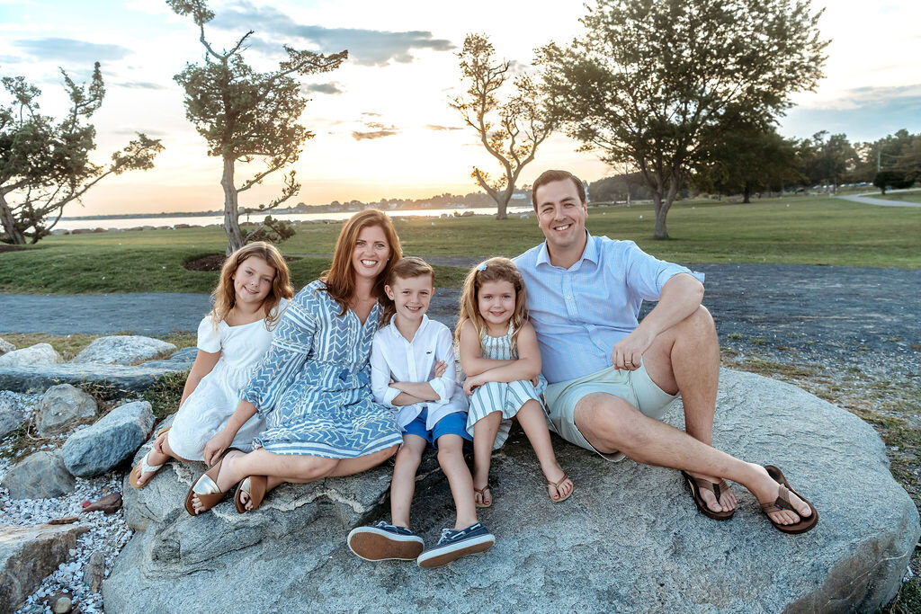 Connecticut family photographer captures family of five sitting on rocks during golden hour beach session with waterfront backdrop