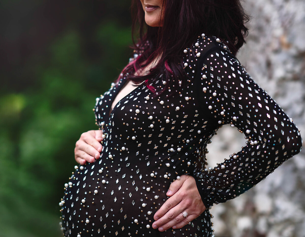 Expectant mom in black rhinestone-embellished maternity gown is standing side profile to the camera. One hand is resting on her hip, the other atop of her bump. Image of of just mom's belly. Captured by Main Line photographer Liza Nicole.