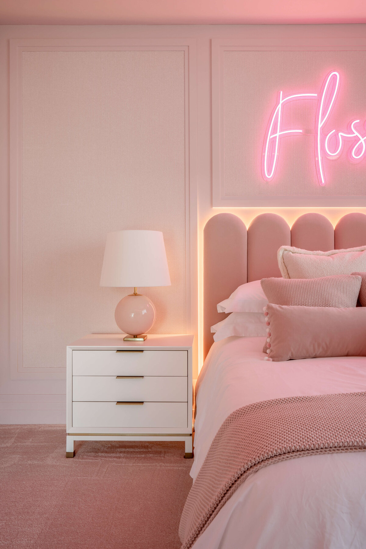 Young girl's room with a pink scalloped velvet headboard, pink bedding, soft pink carpet, a white bedside table. There is a pink neon name sign above the bed.