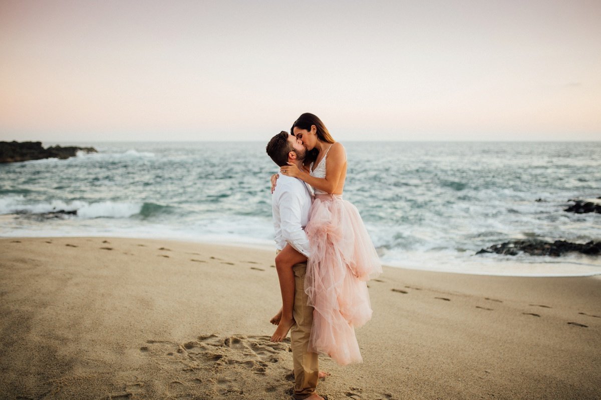 Groom to be lifts his Bride up as she leans down and kisses him during a photo shoot along the beach