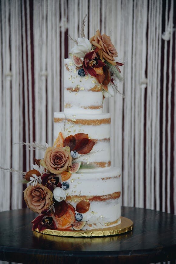 A three tier cake sits on s a cocktail table in front of a macrame curtain. The cake has naked style frosting, with some of the cake showing through, and is decorated with scattered gold leaf flakes and two large clusters of fresh fall flowers and candied fruits.