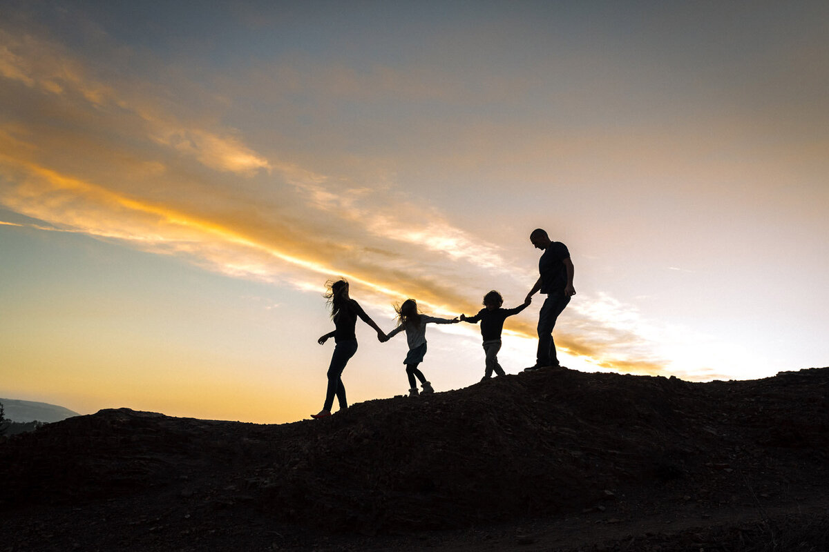 silhouette of family of 4 holding hands and walking together up on a hill at sunset
