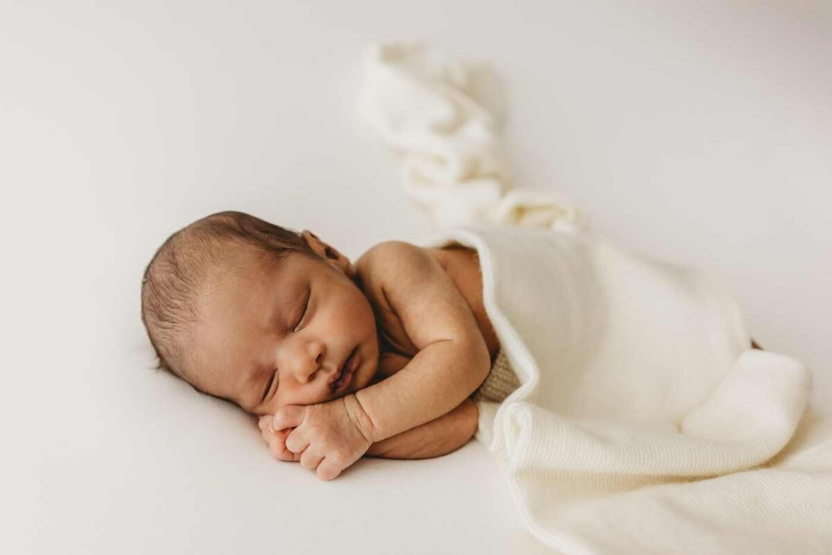 Ruua Hamdan Photography (UK): A sleeping newborn baby is lying on its side, wrapped in a white blanket, and is being held in two pairs of hands.