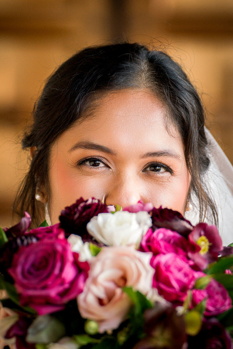 The bride elegantly holds her bouquet in front of her at Casa Real, her eyes peeking over the flowers with a captivating gaze that perfectly balances beauty and mystery on her wedding day.