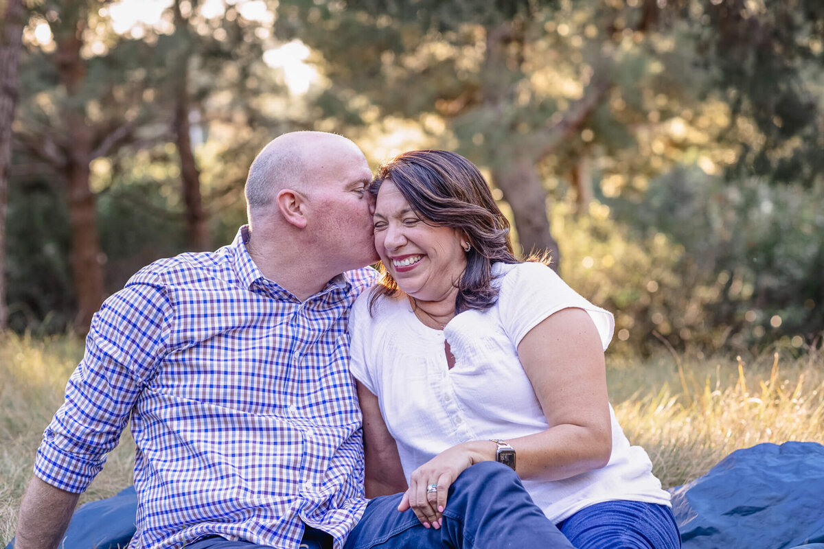 A man wearing a blue plaid dress shirt kissing his wife with short, brown hair wearing a white blouse, sitting on a navy picnic blanket in the grass with trees behind them. Captured at Jeffrey Open Space in Irvine.