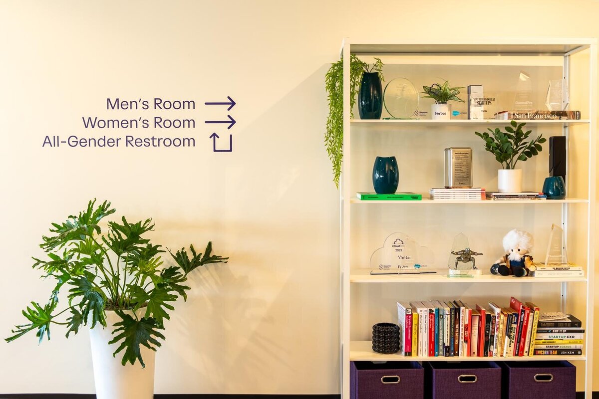Hallway with directional restroom signage and a styled shelving unit displaying awards, books, and plants.