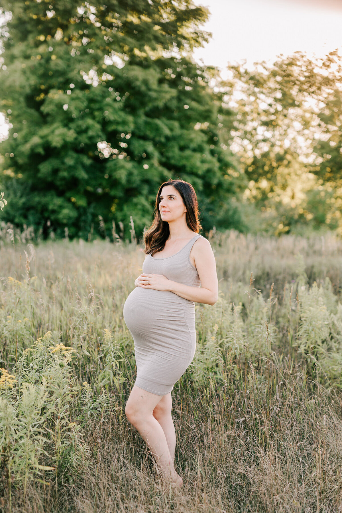 woman in taupe dress smiles at the camera holding her belly.