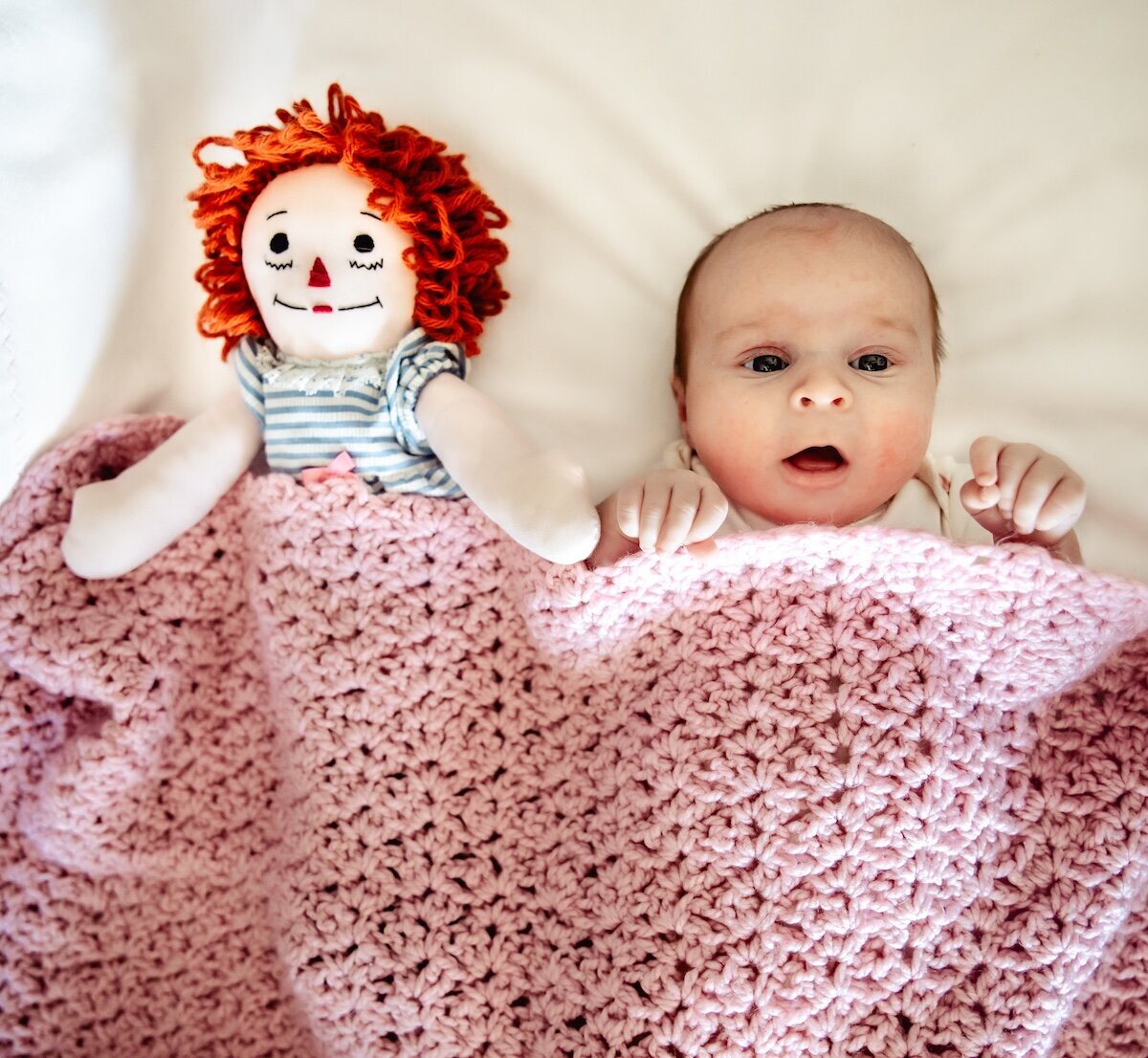 A newborn baby tucked in under a pink knitted blanket next to a stuffed doll