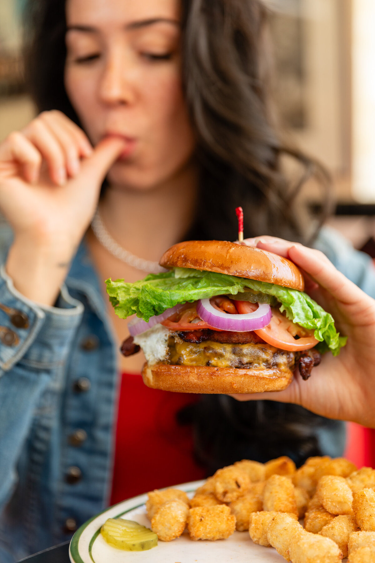 Attractive woman licking sauce off of her finger while holding a fried egg burger in a restaurant.
