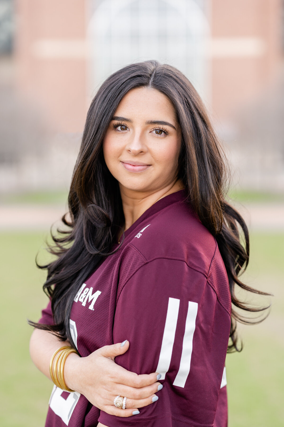 Texas A&M senior girl wearing maroon jersey and smiling over shoulder while holding elbow in Aggie Park in front of Kyle Field