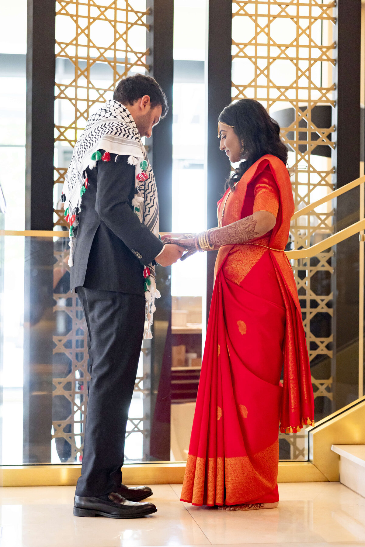 A woman dressed in a vibrant red and orange sari showcases her intricately henna-decorated hands and gold bangles. She wears traditional gold jewelry around her neck and ears, adding to her festive attire.